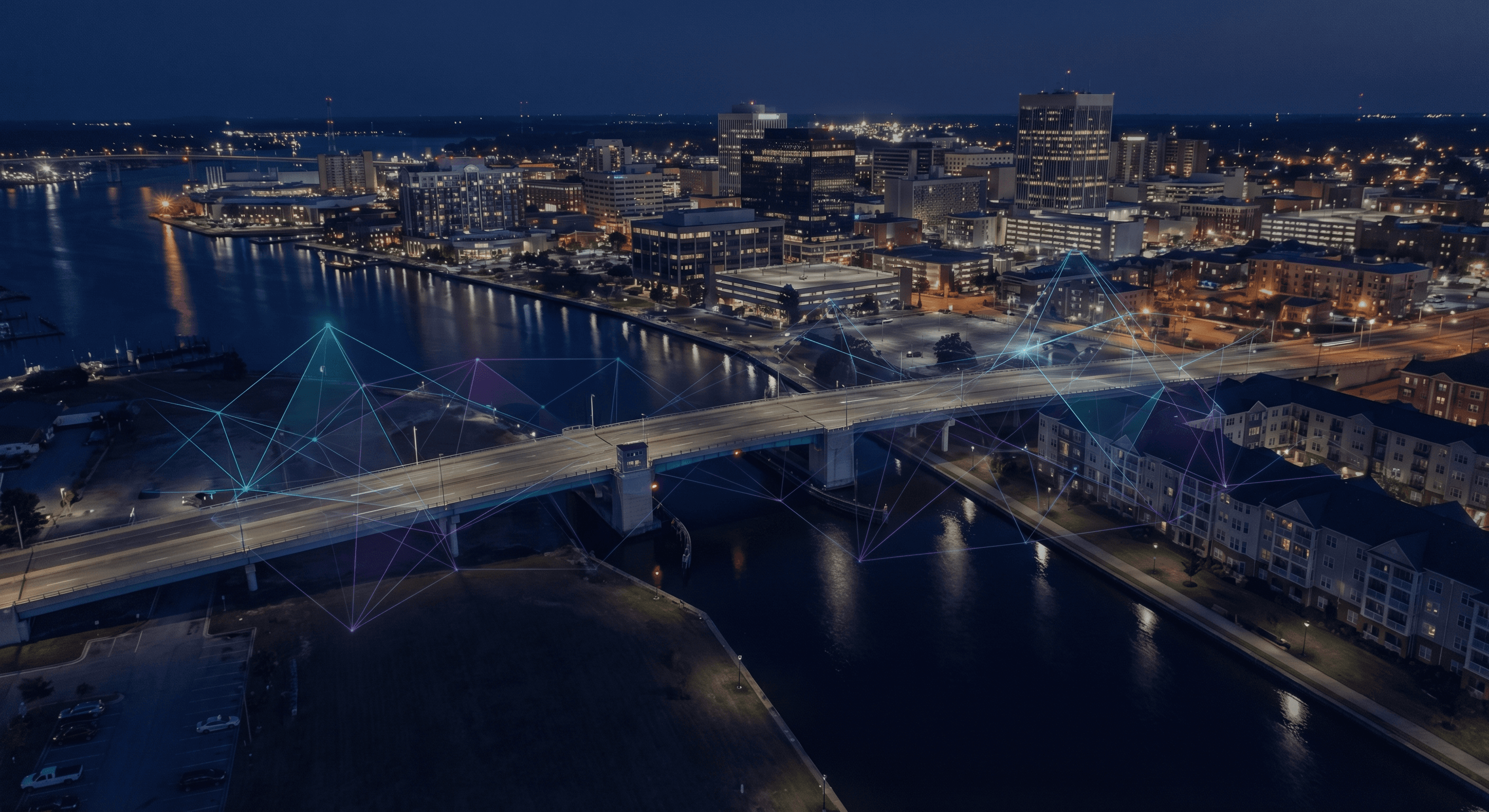 Nighttime aerial view of a waterfront city with a wide bridge crossing a canal, illuminated mid-rise buildings along the shoreline, and reflections in the dark water. Subtle cyan and purple holographic network lines overlay the bridge, streets, and waterfront buildings.
